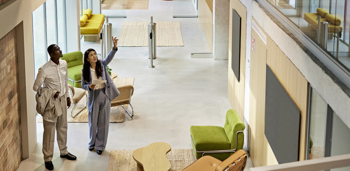 Stock photo for illustration: A black man and Asian woman are standing in the lobby of a two-story, open office space. They're dressed in business clothes and are looking up at 2nd floor space, visible from the lobby. The woman is gesturing as if pointing out features. It appears to be a commercial real estate agent showing a client space for sale or rent.