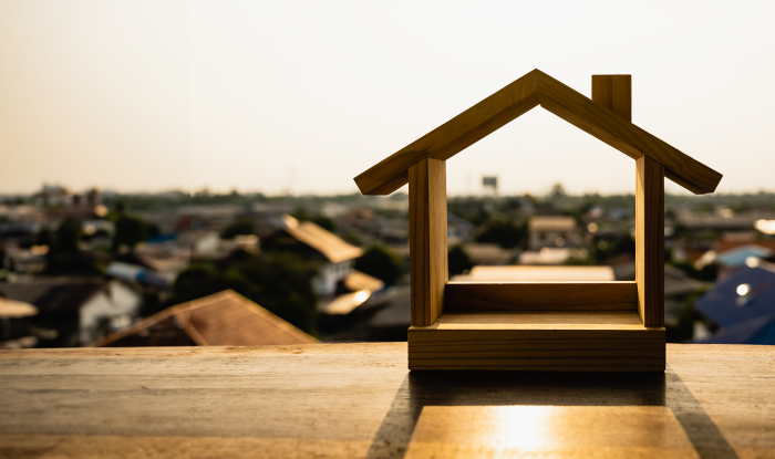 Stock photo for illustration: Wood house frame shape sitting on wooden surface. Rooftops and sunlight can be seen in the background around and through the shape.
